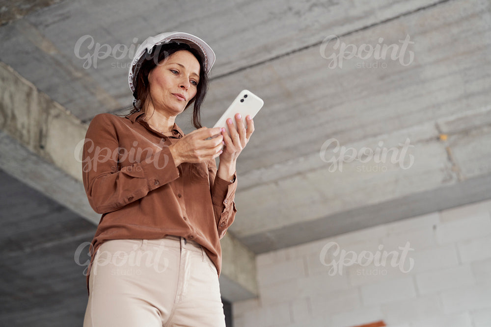 Female mature caucasian engineer standing on construction site and checking something on mobile phone 