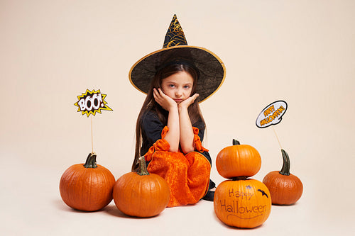 Portrait of bored little witch sitting among halloween pumpkins