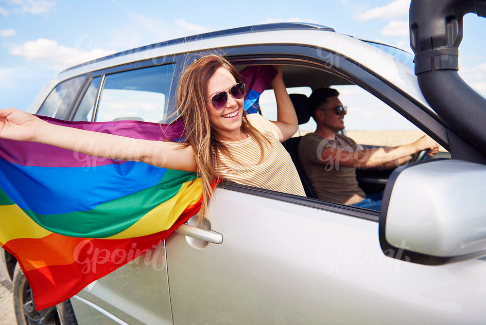 Smiling woman with rainbow flag traveling by car in summertime