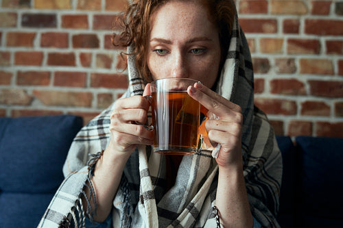 Close up of ill caucasian young woman covered with blanket and drinking hot tea