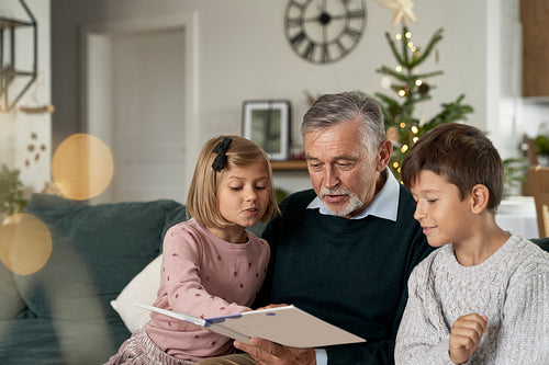 Grandfather and grandchildren reading book on sofa at Christmas time