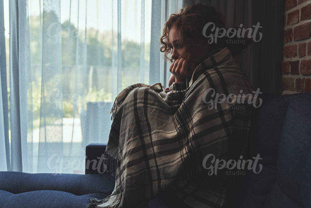Depressed young caucasian woman sitting on sofa in silence covered with blanket 