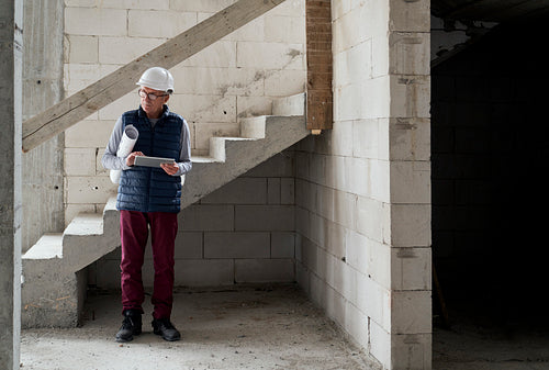 Caucasian senior engineer holding plans and digital tablet while standing on construction site
