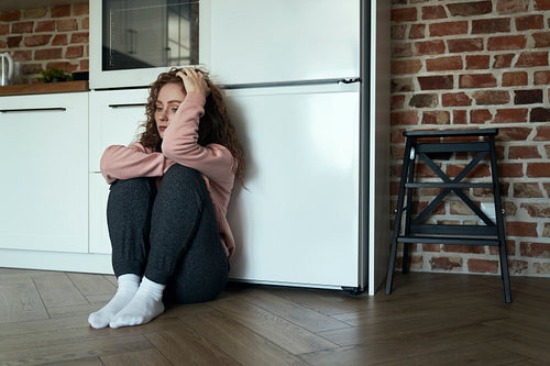 Sad and thoughtful young caucasian woman sitting on floor in the kitchen