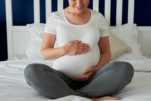 Pregnant woman sitting on bed and keeping hands on stomach