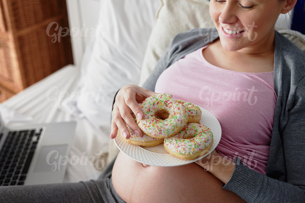 Pregnant woman eating colorful donuts in bed
