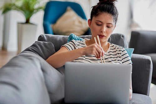 Young woman eating something with sticks in front of laptop.