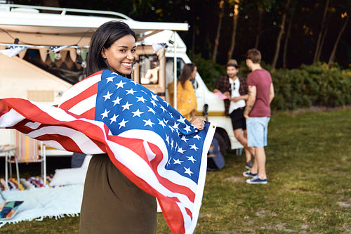 Young mixed race woman covered with USA flag and her friends celebrating 4th of July on the camping