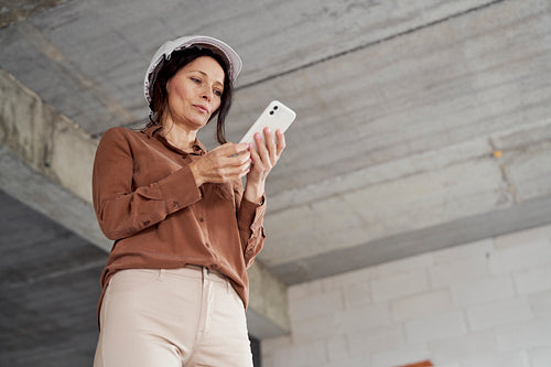 Female mature caucasian engineer standing on construction site and checking something on mobile phone