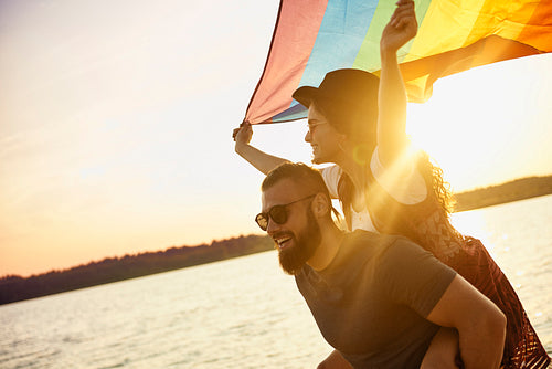 Happy man carrying woman with rainbow flag by sea