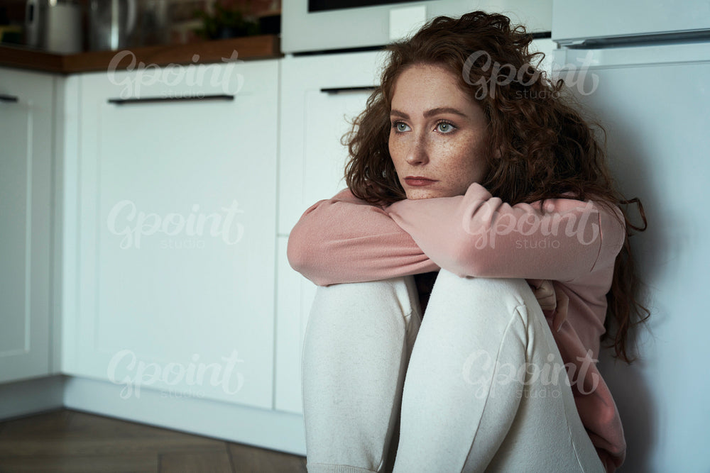 Thoughtful young caucasian woman sitting on floor in the kitchen and sadly looking away