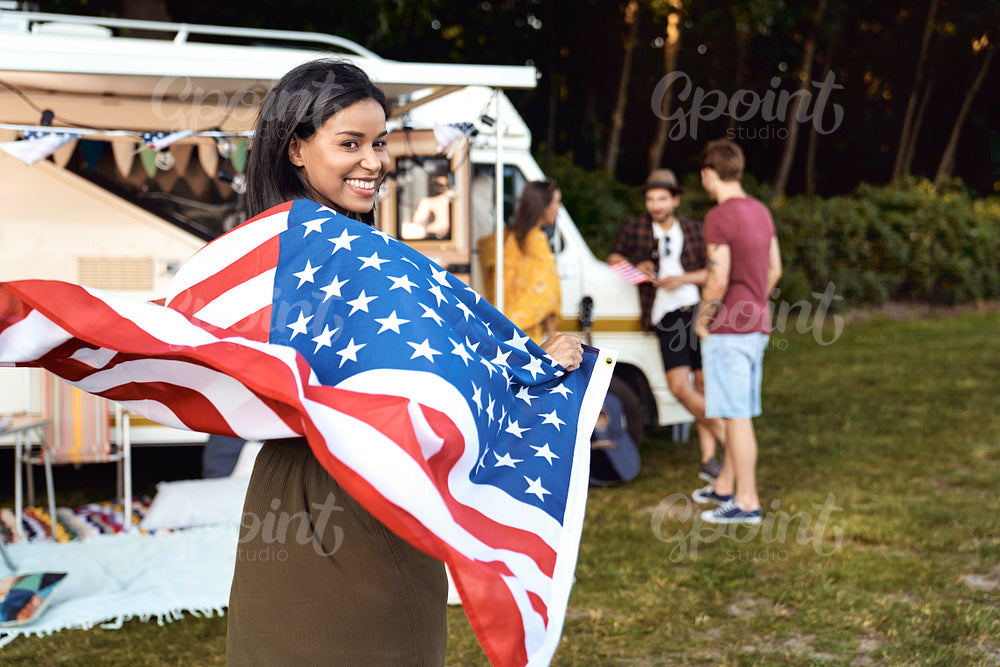 Young mixed race woman covered with USA flag and her friends celebrating 4th of July on the camping 
