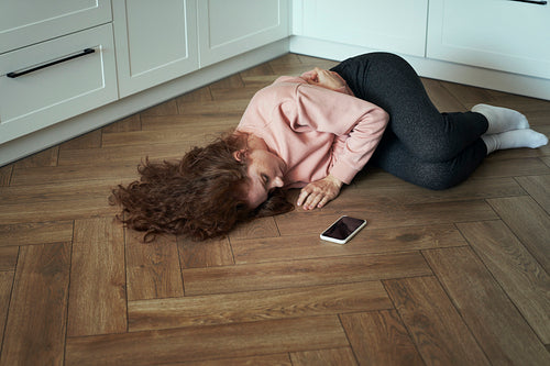 Sad young caucasian woman lying on floor next to mobile phone.