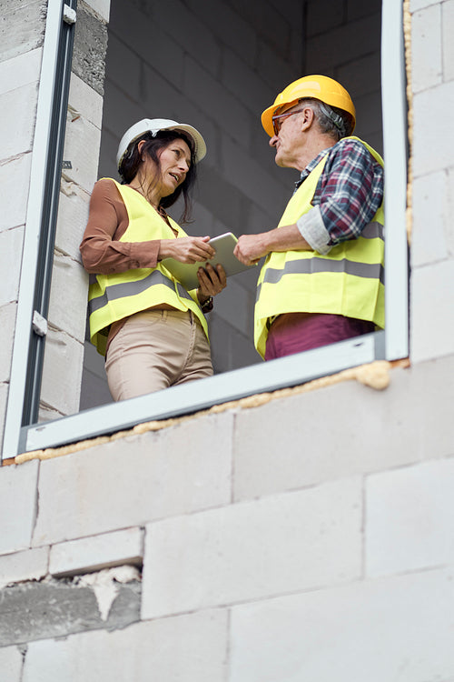 Two caucasian engineers standing near to window and discussing over digital tablet on the construction site