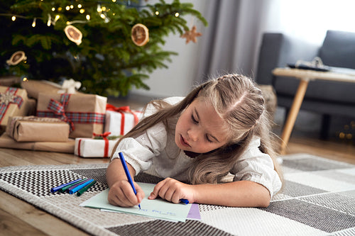 Little caucasian girl lying on carpet next to Christmas tree and writing a letter to Santa Claus