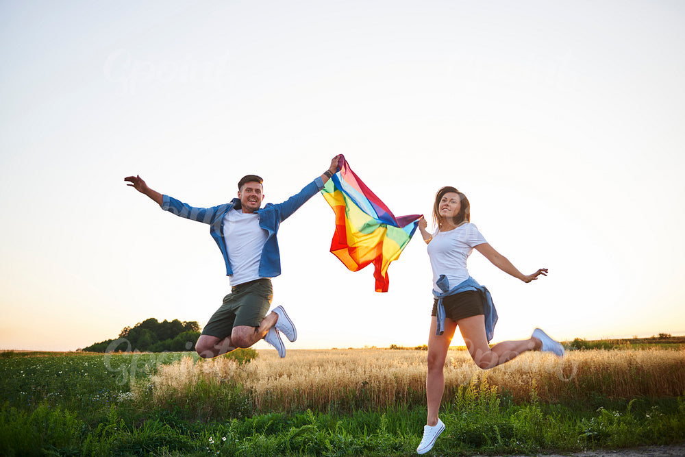 Smiling couple jumping with rainbow flag outdoors.