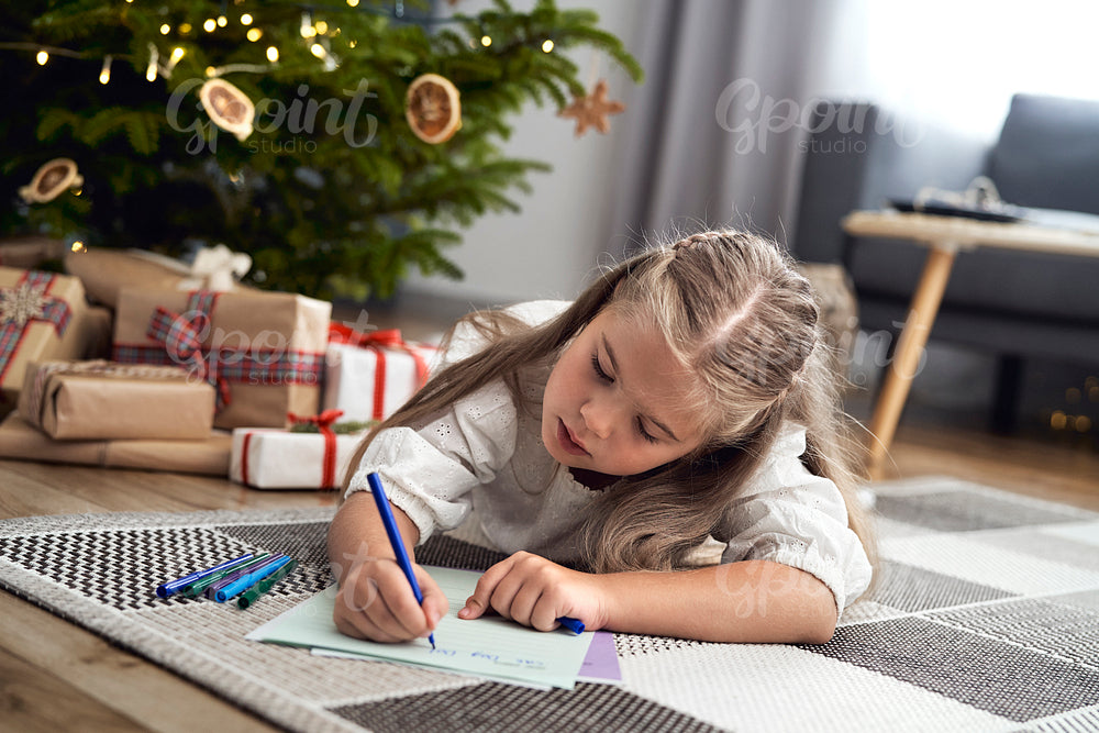 Little caucasian girl lying on carpet next to Christmas tree and writing a letter to Santa Claus