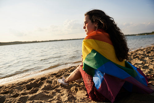 Girl sitting on the beach and watching sunset with rainbow flag