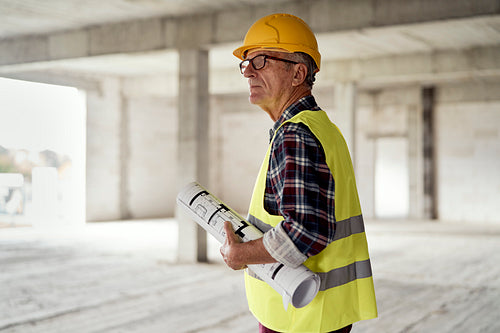 Caucasian senior engineer walking and holding plans on construction site