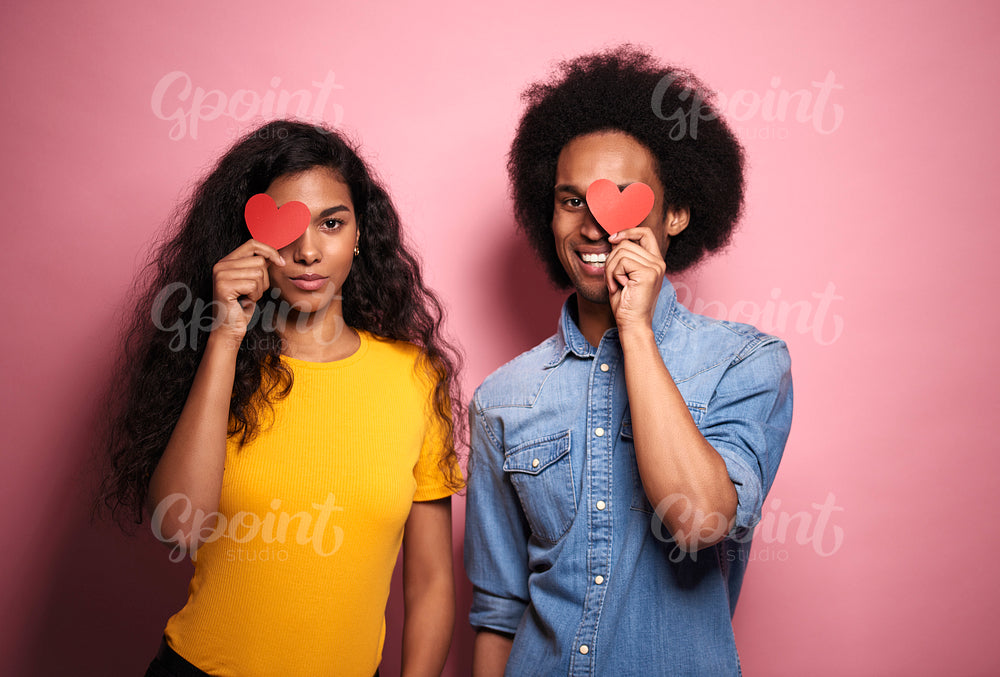 Young African couple covering eyes by hearts in studio shot.