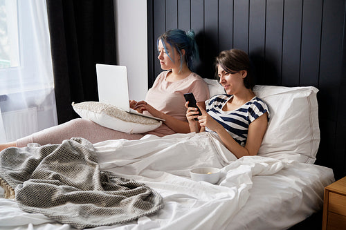 Lesbian couple spending morning in bed with laptop and phone