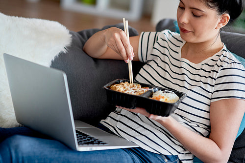 Young Asian woman eating while watching something on a laptop.
