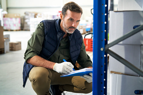 Crouching caucasian men in mature age checking documents and goods in warehouse
